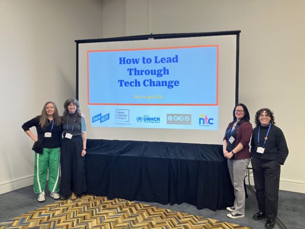 Four people standing on either side of a large screen upon which is projected a title slide for How to Lead Through Tech Change.